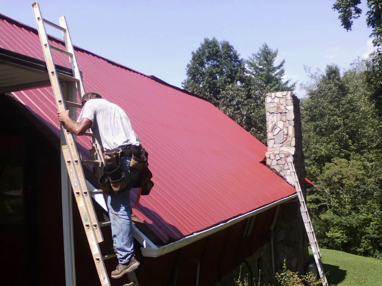 Roofer on a ladder beside a red metal roof and stone chimney — Soffit & Fascia Repair by Roofing Brothers in East Helena